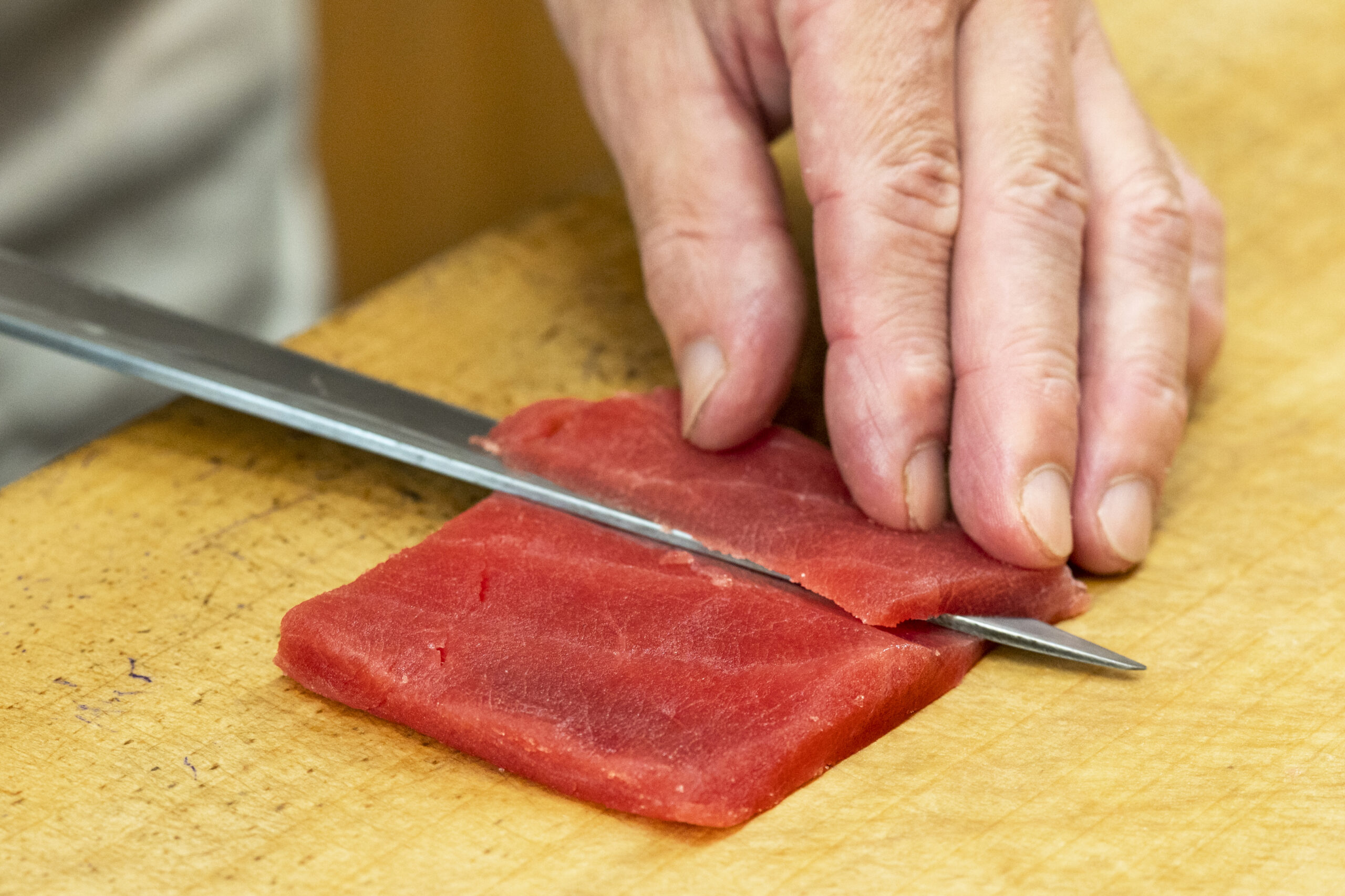 Hands slicing a block of tuna with a long knife on a wooden cutting board
