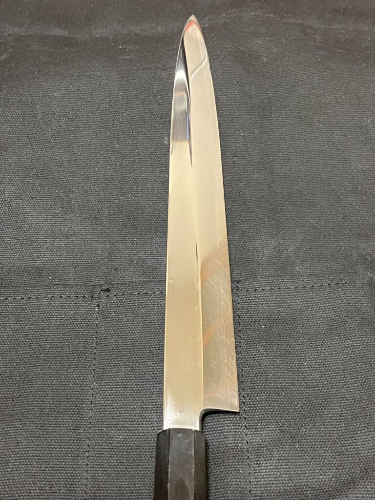 Close-up of a long, narrow yanagiba knife on a dark textured surface